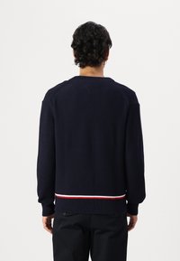 Marineblå ribbestrikket genser med lange ermer og rund hals. Har en horisontal stripe i rødt og hvitt ved kanten. Teksturert strikket stoff.