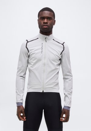 Mężczyzna ubrany w jasnoszarą kurtkę zapinaną na zamek z czarnymi akcentami i czarne spodnie sportowe, stojący na jednolitym białym tle.