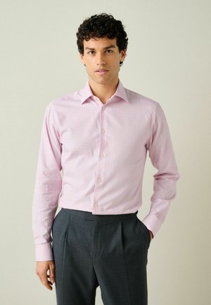Man wearing a light pink checkered long-sleeve button-up shirt tucked into dark gray pleated trousers, standing with one hand in pocket.