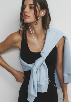 Melns tanktops kombinācijā ar gaiši zilu džemperi, kas ir sasiets viduklī. Modelim ir baltas austiņas un viegli pajukuši mati. Vienkāršs fons.