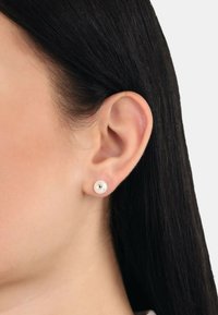 Runda, vita studs örhängen med en liten guldfärgad hjärtan detalj i mitten, mot en bakgrund av rakt, mörkt hår.