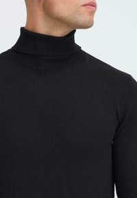 Hombre vistiendo un suéter de cuello alto acanalado negro, mostrado desde el cuello hasta la parte superior del pecho, de perfil a la derecha contra un fondo sencillo.
