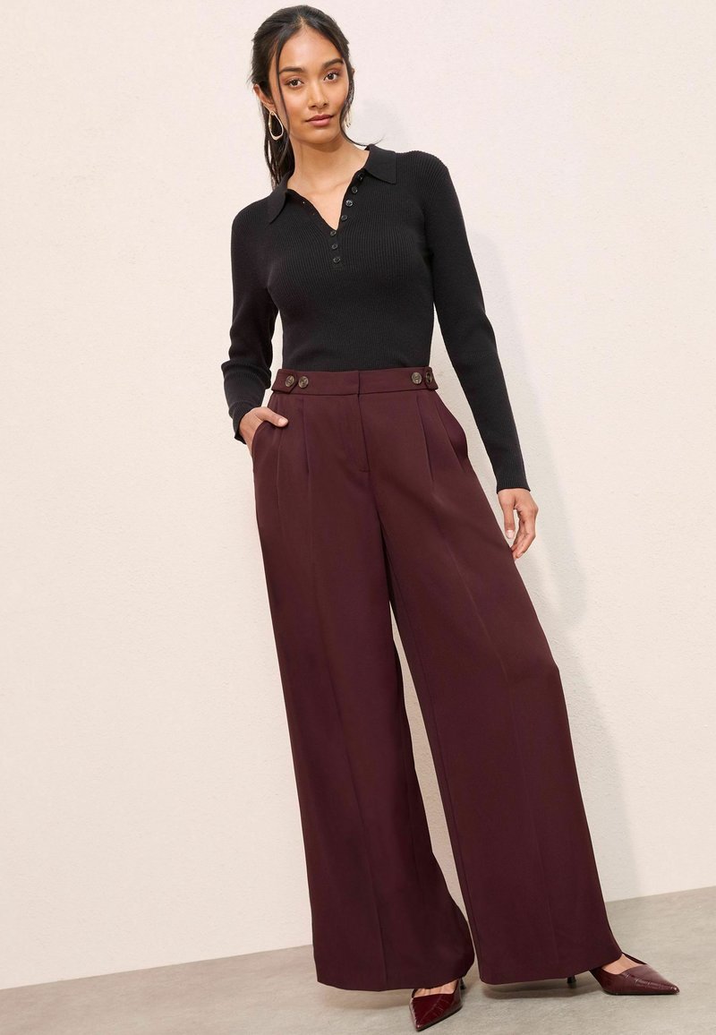 Črn rebrast longsleeve top z ovratnikom in gumbnim zapenjanjem, v kombinaciji z širokimi burgundskimi hlačami, ki imajo sprednje nabore in gumbne detajle.