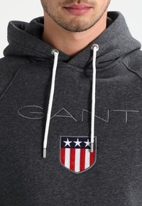 Hoodie grigio scuro con coulisse bianche, toppa ricamata a forma di scudo con tre stelle bianche sopra strisce rosse e bianche, e logo "GANT" sul petto.