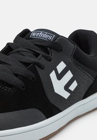 Μαύρα αθλητικά παπούτσια που συνδυάζουν σουέτ και διχτυωτά υλικά, με λευκό λογότυπο, υφασμάτινες λεπτομέρειες και λευκή καουτσούκ σόλα.