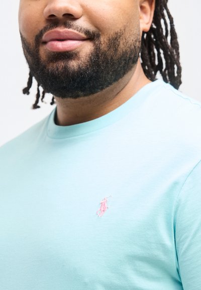 Άντρας με γενειάδα και κοτσίδες φορώντας ανοιχτό μπλε πουκάμισο με μικρό ροζ λογότυπο παίκτη πόλο στο στήθος σε λευκό φόντο.