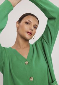 Zielony kardigan wykonany z miękkiej dzianiny, z dekoltem w kształcie V, długimi rękawami i dwoma dużymi guzikami. Gładka tekstura, luźny krój i kieszenie.