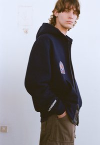 Tmavomodrá kapucňová mikina na zips s nášivkou s logom na hrudi, klokanie vrecko a nápis "LAND CRUISER" na rukáve. Nosená s olivovo zelenými nohavicami.