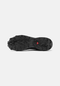 Semelle de chaussure de sport noire avec des rainures en caoutchouc prononcées et un accent rouge. Surface texturée conçue pour une traction sur divers terrains.