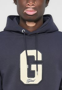 Marineblå hættetrøje med et stort cremefarvet fleece "G" logo og en sort omridset hexagonal kant. Har snoretræk og en blød tekstur.