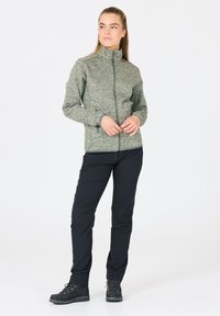 Kvinna som står iförd en grön zip-up jacka, svarta byxor och svarta vandringskängor, tittar bort mot sidan med händerna hopknäppta.