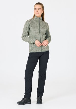 Grön texturerad zip-up jacka med ståkrage, kombinerad med svarta byxor. Skorna har svarta detaljer och robusta sula.