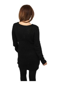 Donna con capelli castano medio, che indossa un lungo maglione nero lavorato a maglia e pantaloni neri, di spalle su uno sfondo bianco.