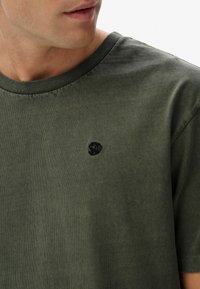 Grøn bomulds t-shirt med rund halsudskæring, korte ærmer og et lille sort broderet logo på brystet. Blødt, struktureret stof.