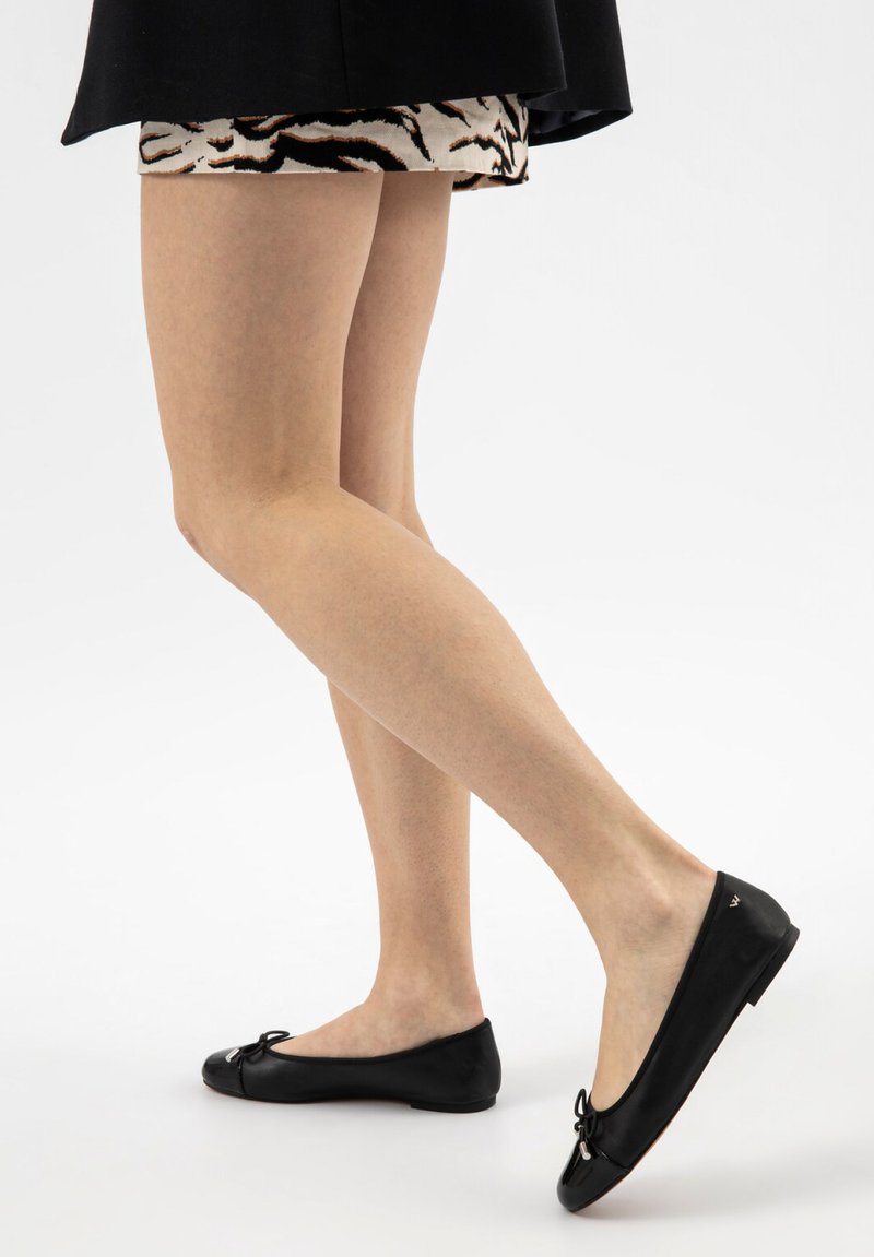 Žena ve černé sukni a šatech se vzorem zvířecí kůže si nazouvá černé kožené baleríny s malými mašličkami.