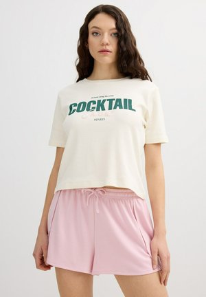 Mujer con el pelo largo y castaño que lleva una camiseta blanca con la palabra "COCKTAIL" y pantalones cortos rosa claro sobre un fondo blanco liso.
