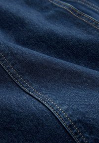 Acercamiento de una tela de mezclilla azul oscuro con costuras naranjas visibles a lo largo de las costuras curvadas.