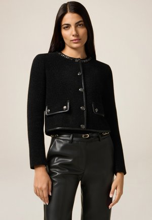 Zwart cropped jack met metalen knopen, textuurstof en leren afwerking. Beschikt over twee voorkanten zakken en een decoratieve kraagdetail.