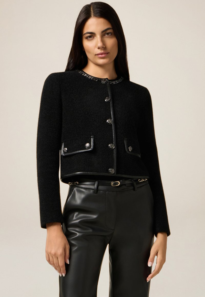 Sort cropped jakke med metallic knapper, struktureret stof og læderdetaljer. Har to forlommer og en dekorativ kravedetalje.
