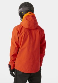 Chaqueta naranja impermeable con capucha, con puños negros y un logo de HH en el cuello. Textura suave, diseñada para actividades al aire libre.