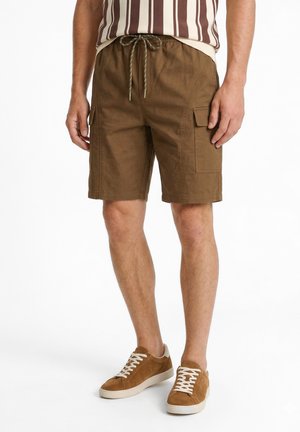 Personne portant un short cargo marron avec taille à cordon, un t-shirt rayé et des baskets en daim marron avec des lacets blancs, debout sur fond blanc.
