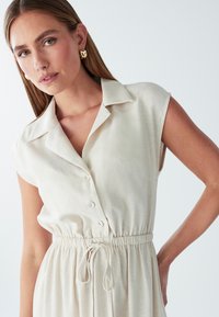 Beež, lühikeste varrukatega nööpidega kleit krae ja elastse vöökohtaga, millel on lips. Pehme, tekstuuriga kangas koos voolujoonelise disainiga.