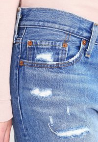 Světle modré denimové džíny s odřeným designem, třepenými okraji a několika malými dírami. Jsou viditelné detaily z mědi.