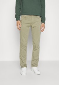 Man som bär beige chinos, grön sweatshirt och vita sneakers, ståendes mot en enkel ljus färgad vägg.