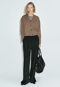 Cardigan marrone, texturizzato, con bottoni, abbinato a pantaloni neri a vita alta. Borsa in pelle nera, posizionata su uno sfondo neutro.