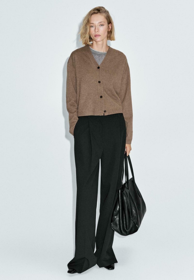 Cardigan marrone, texturizzato, con bottoni, abbinato a pantaloni neri a vita alta. Borsa in pelle nera, posizionata su uno sfondo neutro.