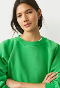 Zaļš džemperis, kas izgatavots no mīksta auduma, ar apaļu kakla izgriezumu un raglāna piedurknēm. Tekstūra šķiet gluda un tas ir brīvi piegriezts.