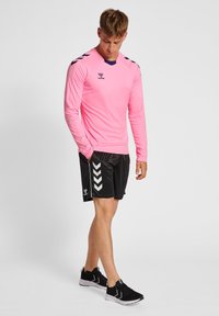 Maglietta atletica rosa a maniche lunghe con colletto viola, abbinata a pantaloni corti neri con dettagli bianchi a chevron. Scarpe da ginnastica nere.