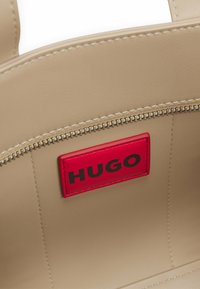 Beige handbag featuring a red leather label with "HUGO" in black. The zip closure is accentuated with metallic hardware. Smooth texture.