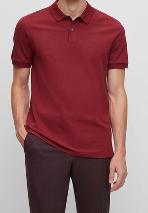 Mann trägt ein dunkelrotes Poloshirt mit kurzen Ärmeln und Kragen sowie dunkle Hosen, steht mit einer Hand in der Tasche.