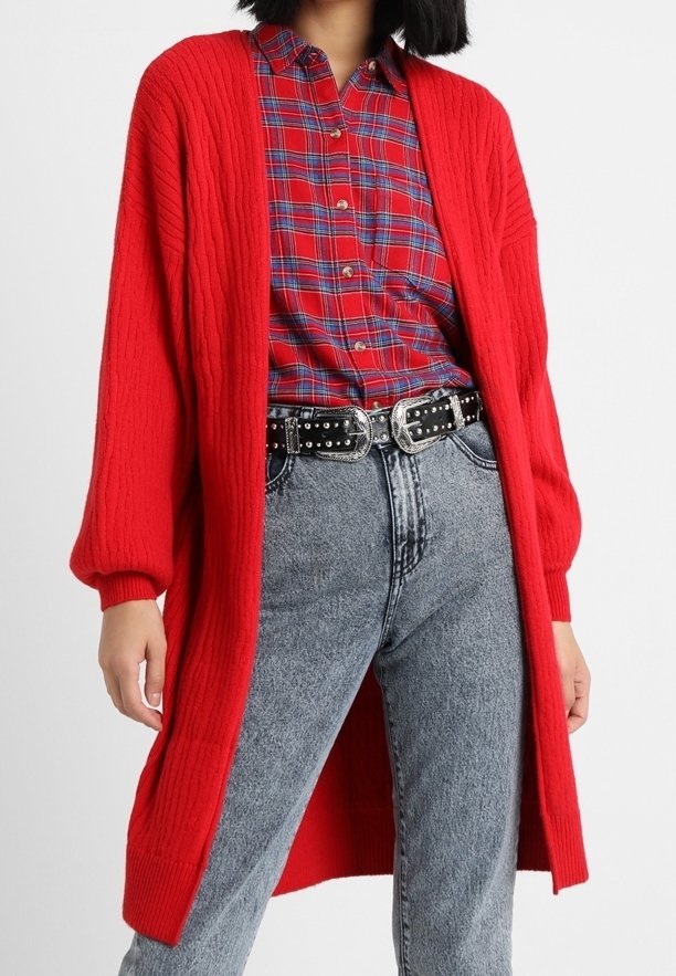 Rode gebreide cardigan met lange mouwen, geribbelde textuur en open voorkant, gelaagd over een rood-blauw geruit shirt en hooggetailleerde jeans.