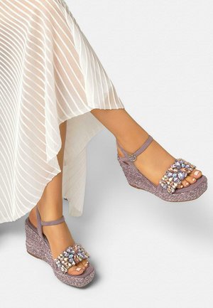 Benen in een witte, vloeiende geplooide rok, met lavendelkleurige sleehaksandalen met enkelbandjes en meerkleurige juweelversieringen.