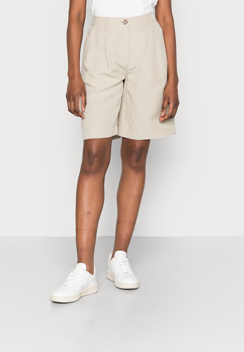Personne portant un short beige longueur genoux, des baskets blanches et un haut blanc sur un fond uni clair.