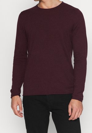 Uomo che indossa un maglione a maniche lunghe color borgogna e pantaloni neri, in piedi contro uno sfondo chiaro e semplice.