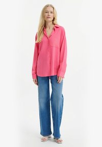Světle růžová oversized košile na knoflíky s kapsou na prsou, kombinovaná s širokými modrými džíny a bílými sandály na podpatku.