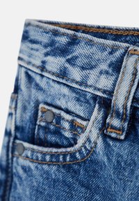 Los jeans de denim presentan un primer plano de una tela azul deslavada con costuras visibles y dos botones metálicos, mostrando un bolsillo delantero y la cinturilla.