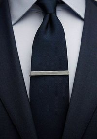 Silverfärgad slipsklämma med horisontella spår som säkrar en mörk marinblå slips. Slipsen har en slät, texturerad tyg mot en ljusblå skjorta.
