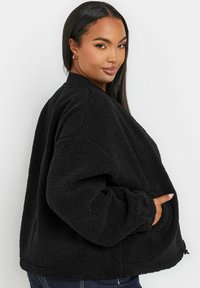 Czarna polarowa kurtka z teksturowaną powierzchnią, prążkowanym kołnierzem i bocznymi kieszeniami, o luźnym kroju i szerokich rękawach.