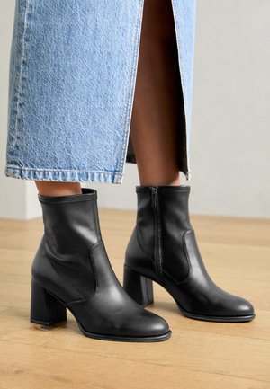 Bottes noires en cuir à talons carrés portées avec une jupe en jean bleu clair avec une fente devant, debout sur un sol en bois.