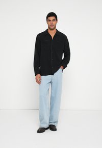 Musta nappipaita, jossa on kaksi rintataskua, yhdistetty vaaleansinisiin leveälahkeisiin farkkuihin ja mustiin kenkiin, valkoista taustaa vasten.