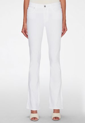 Femme portant un jean bootcut blanc taille haute avec fermeture bouton et zip, associé à des chaussons crème à bouts ouverts.