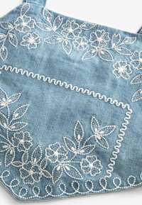 Borsa in denim blu con ricami floreali bianchi, cuciture decorative e una tasca frontale. Tessuto liscio con motivi e dettagli intricati.