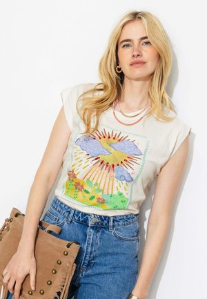 Jeune femme aux longs cheveux blonds portant un top blanc sans manches avec un motif de soleil, nuages et fleurs, un jean bleu et tenant un sac marron à franges.