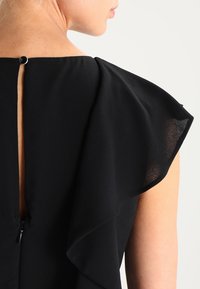 Czarna bluzka z falbanianymi rękawami, z wycięciem na plecach i małym czarnym guzikiem nad ukrytym zamkiem.