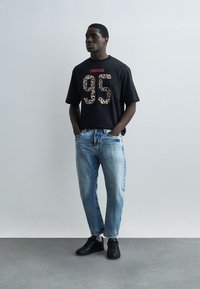 Άνδρας φορώντας μαύρο t-shirt DSQUARED2 με αριθμό 95 σε λεοπάρ μοτίβο, ανοιχτό μπλε τζιν, μαύρα αθλητικά παπούτσια, στέκεται με τα χέρια στις τσέπες.