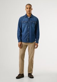 Donkerblauwe denim shirt voor heren met twee borstzakken, knoopsluiting en contrasterende stiksels, gecombineerd met beige corduroypantalon en bruine schoenen.